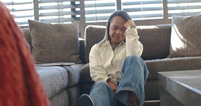 African American woman leaning on sofa in living room, listening to off-frame voice with laptop