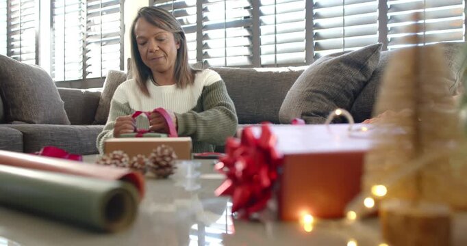 Mature African American woman on couch wrapping kraft box tying pink ribbon into bow preparing gift