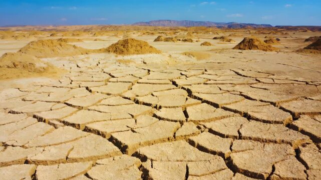 Desert floor cracked wide under blazing sun. Earth's surface split by harsh, dry weather. Dust and rock dominate this barren expanse. Vast, empty landscape stretches to distant hills