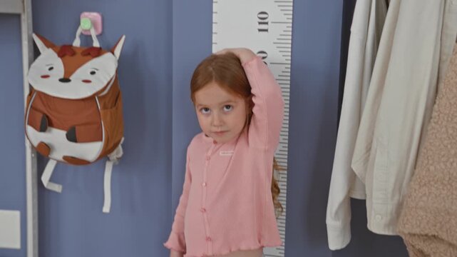 Girl places hand on head while measuring height against wall growth chart in building; growth milestone curiosity.