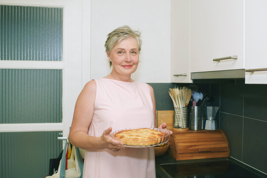 55-60 year old woman holding fresh homemade pie in modern kitchen, smiling mature female enjoying baking and preparing delicious dessert at home
