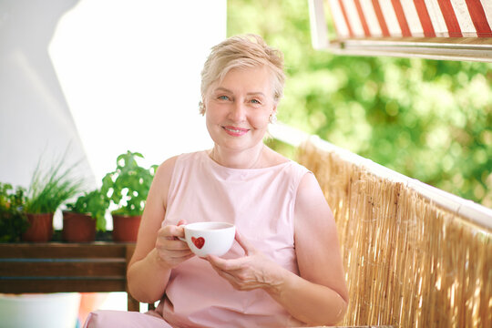 55-60 year old woman holding a cup with red heart, smiling and enjoying a warm drink on sunny balcony, concept of love, happiness and relationship