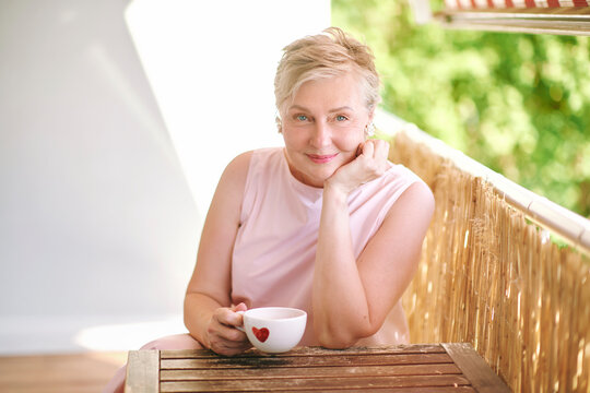 55-60 year old woman holding a cup with red heart and smiling on sunny balcony, happy mature female enjoying a warm drink, love and relationship concept