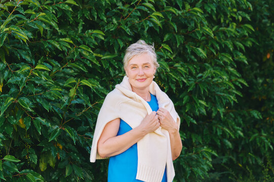 55-60 year old woman smiling outdoors in green garden in autumn, confident mature female wearing blue dress and warm sweater, enjoying nature and peaceful lifestyle
