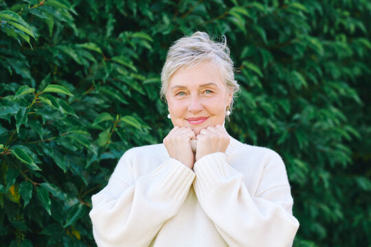 55-60 year old woman wearing warm white sweater outdoors in autumn, smiling mature female portrait surrounded by green leaves, cozy seasonal lifestyle and natural beauty