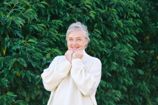 55-60 year old woman wearing warm white sweater outdoors in autumn, smiling mature female portrait surrounded by green leaves, cozy seasonal lifestyle and natural beauty