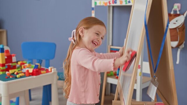 Girl erases whiteboard on easel with hand using red eraser in a building classroom with blocks and toys nearby; learning concentration.