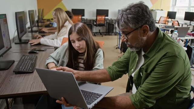 Middle aged South Asian male teacher explaining coding on laptop to Caucasian female teenager in computer lab, high angle shot. Perfect for promoting STEM education and digital literacy programs