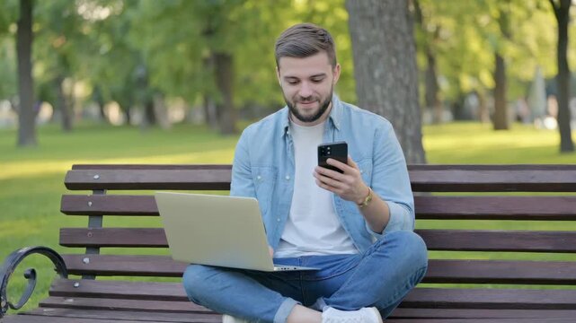 Man sits park bench using laptop and phone. Trees and grass surround him in green park. He wears jeans, shirt, and white sneakers. Distant walker moves through blurred background