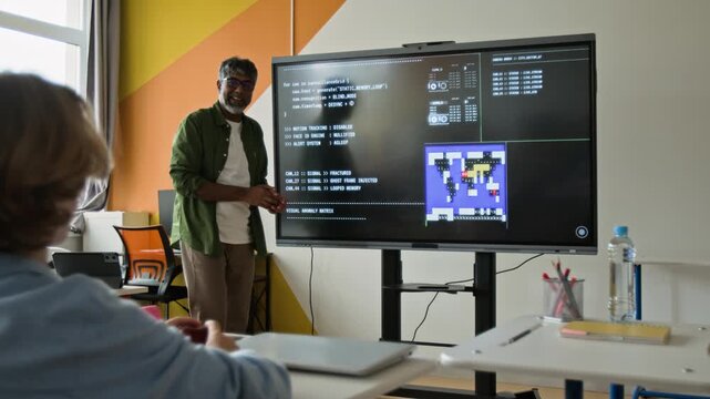 Indian middle aged teacher explaining programming code on screen to Caucasian teenager seen from rear view raising hand. Perfect for STEM education and coding school marketing campaigns