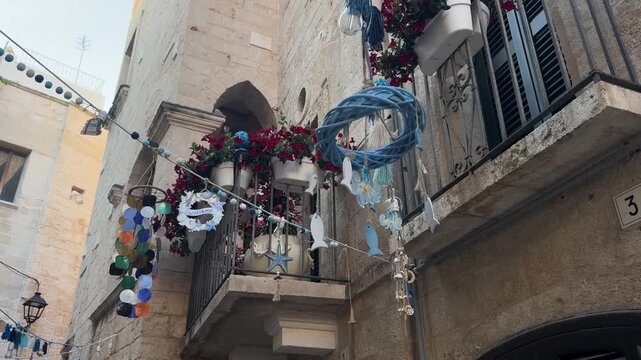 Traditional stone buildings in Monopoli old town, Italy, feature a balcony with hanging ornaments and street decorations beneath a clear blue sky. Puglia travel scene in a historic European town area.