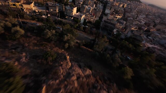 Dynamic fast-paced FPV drone footage diving past residential rooftops and apartment buildings in an urban Athens neighborhood facing Lycabettus Hill and the warm golden flare of sunset.