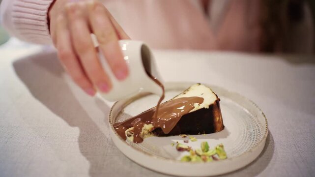 Woman pouring warm liquid chocolate sauce over slice of gourmet basque cheesecake on plate.