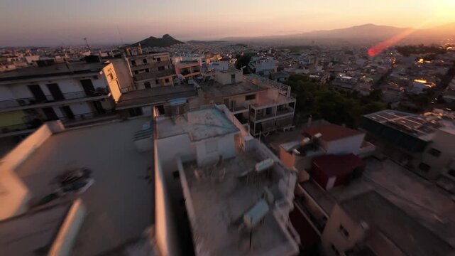 Dynamic fast-paced FPV drone footage diving past residential rooftops and apartment buildings in an urban Athens neighborhood facing Lycabettus Hill and the warm golden flare of sunset.