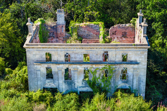 Ruins of 18th century Piercefield House in Chepstow, roofless stone facade and brick chimneys surrounded by dense green summer woodland, high angle aerial view