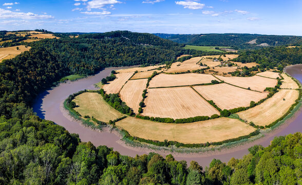 High angle panoramic view of river curving around yellow harvest fields, dense green woodland covering valley hillsides on sunny summer afternoon