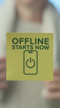 Close up of hands placing a yellow sticky note on a glass surface, the message offline starts now with a smartphone power off icon, encouraging a break from digital technology and social media