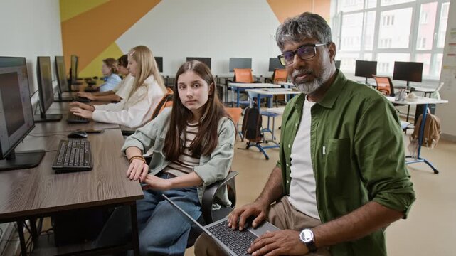 Portrait of South Asian middle aged male teacher and Caucasian female teenager looking at camera in computer class. Footage is ideal for promoting STEM education and coding bootcamps