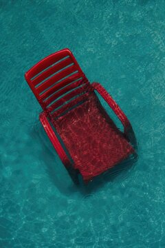 Red plastic chair partially submerged in clear blue swimming pool water, minimalist modern design, abstract reflection, leisure and vacation concept