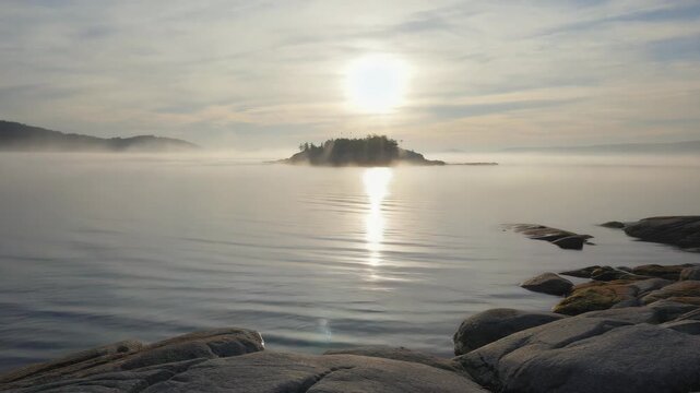 Early morning sun reflecting on calm water, with fog and a distant island.