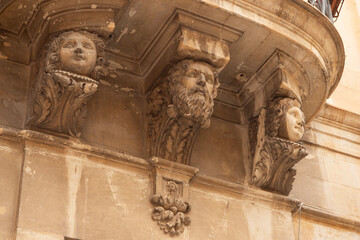Modica, Italy  1st August 2026: Baroque architectural detail with carved grotesque face and ornate stonework on a balcony in Sicily. © Leckerstudio