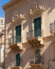 Modica, Italy  1st August 2026: Baroque architectural detail with carved grotesque face and ornate stonework on a balcony in Sicily. © Leckerstudio