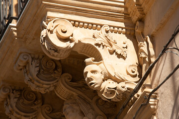 Modica, Italy  1st August 2026: Baroque architectural detail with carved grotesque face and ornate stonework on a balcony in Sicily. © Leckerstudio