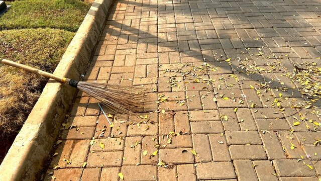 A traditional outdoor broom lying on a paved brick walkway while scattered yellow leaves are being swept. The scene represents outdoor cleaning, maintenance and everyday urban landscape care