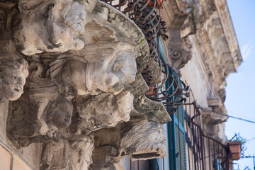 Modica, Italy  1st August 2026: Baroque architectural detail with carved grotesque face and ornate stonework on a balcony in Sicily. © Leckerstudio