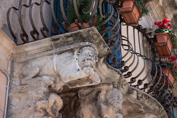 Modica, Italy  1st August 2026: Baroque architectural detail with carved grotesque face and ornate stonework on a balcony in Sicily. © Leckerstudio