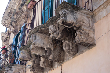 Modica, Italy  1st August 2026: Baroque architectural detail with carved grotesque face and ornate stonework on a balcony in Sicily. © Leckerstudio