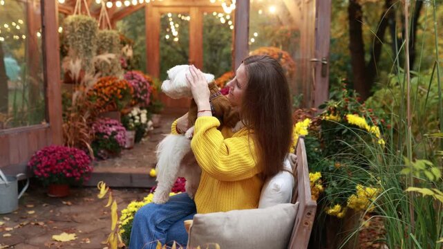 Happy girl pet owner holding a small white dog in a cozy fall garden filled with potted flowers. Warm autumn evening landscape with glowing greenhouse lights, backyard, pet friendship concept.