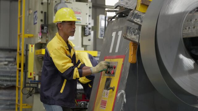 Senior Asian worker wearing yellow hardhat operating control panel near steel coil machine inside factory, technician pressing button industrial manufacturing plant