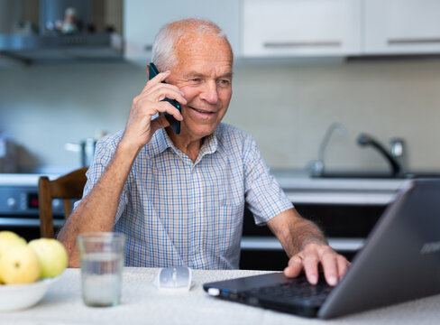 Mature man buys stock online by mobile phone using laptop and internet while sitting in kitchen of his home