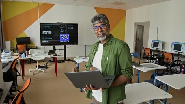 Portrait of South Asian middle aged man holding laptop in computer science classroom, looking at camera and smiling. Video can be used for promoting STEM education and IT training programs