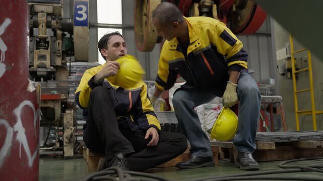 Tired diverse engineers sitting on floor resting after hard work, senior Asian man young Caucasian worker suffering heat exhaustion inside manufacturing factory