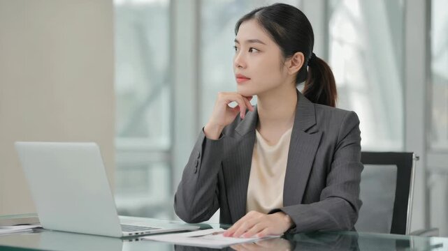 A young woman wearing a suit and sitting at a desk, concentrating on work on her laptop.