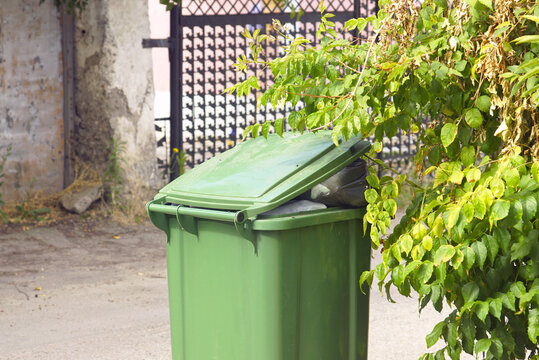 Green wheelie bin outdoors with lid ajar and garbage bags visible inside. Depicts overfilled household waste, urban sanitation strain, and tension between limited capacity and daily refuse generation.