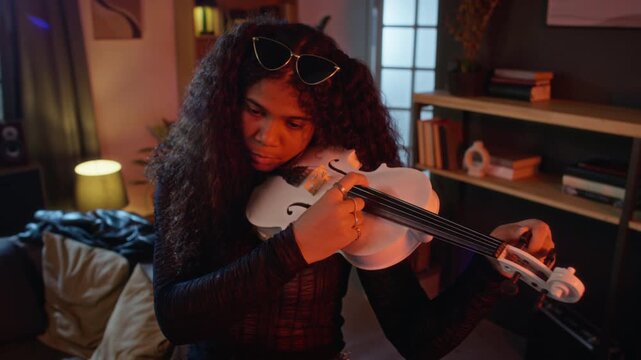 African American teenager tuning electric violin in dimly lit room, adjusting pegs with focus. Footage suitable for music education campaigns or creative arts advertisements