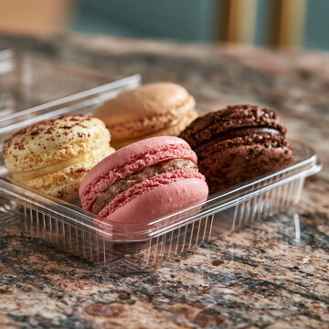 Assorted sweet macarons arranged in a clear plastic container on a table with baked goods