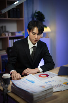 Asian Businessman Working Late Night in Office, Serious Executive Reading Financial Paperwork and Analyzing Reports at Desk