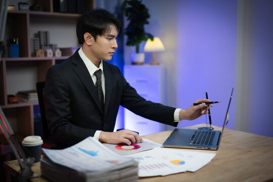Asian Businessman Working Late Night in Office, Serious Executive Reading Financial Paperwork and Analyzing Reports at Desk