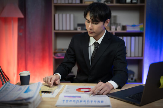 Asian Businessman Working Late Night in Office, Serious Executive Reading Financial Paperwork and Analyzing Reports at Desk