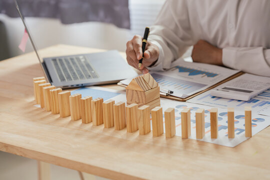 Businessman Analyzing Financial Charts and Calculating Risk Management with Wooden Blocks Domino Line Leading to House Model