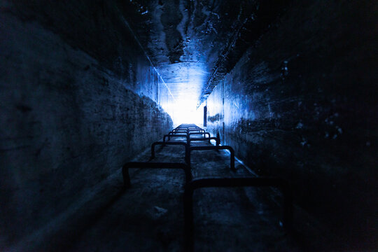 pozzo stretto sotterraneo e buio in un luogo abbandonato, con pareti in cemento e una scala centrale che conduce verso una luce blu brillante in fondo