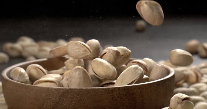 Wooden bowl filled with pistachios on dark wooden table, tasty salted nuts scattered around, healthy snack and natural food concept. Shot on super slow motion camera 1600 fps.