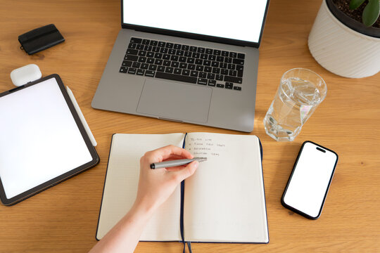 Hand writing a to-do list in a notebook with digital workspace with laptop and tablet and phone, symbolizing business planning, research, creativity and getting things done