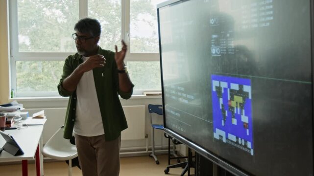 Middle aged South Asian teacher explaining programming concepts on interactive smartboard in classroom. Ideal for coding bootcamp advertisements and educational technology marketing
