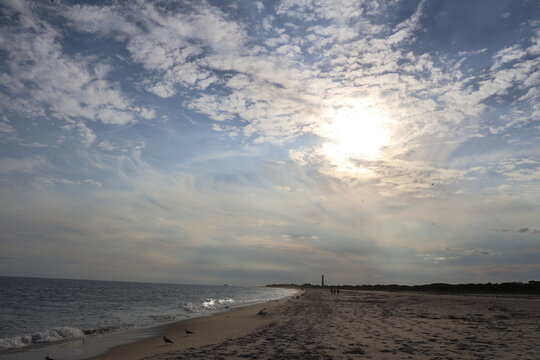 An orange summer sun peaks through a thin layer of clouds and haze onto a peaceful empty beach as the tide gently rolls up on the sand.