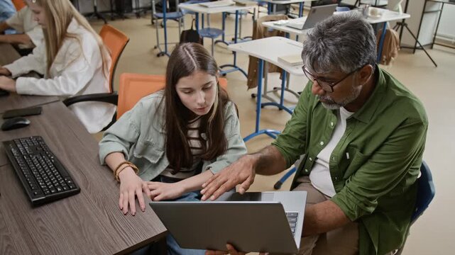 Middle aged South Asian male teacher explaining coding on laptop to Caucasian female teenager in classroom, high angle shot. Footage is perfect for promoting STEM education programs
